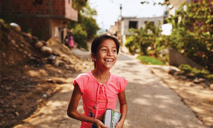 girl smiles boardly as she walks down a villiage street with a Bible under her arm